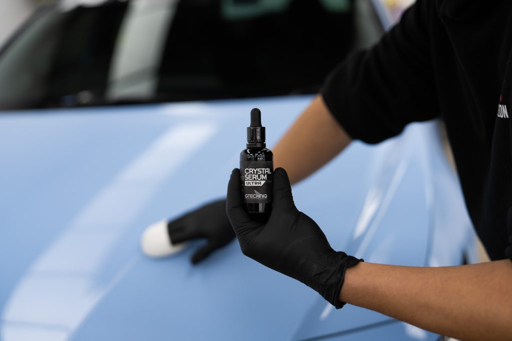 Technician holding Gtechniq Crystal Serum bottle and applicator beside a blue car
